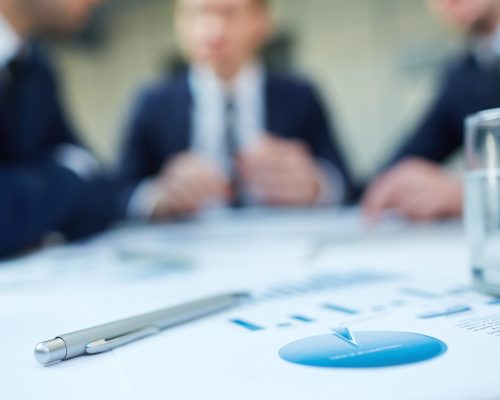 Image of business document and pen at workplace with group of colleagues interacting on background